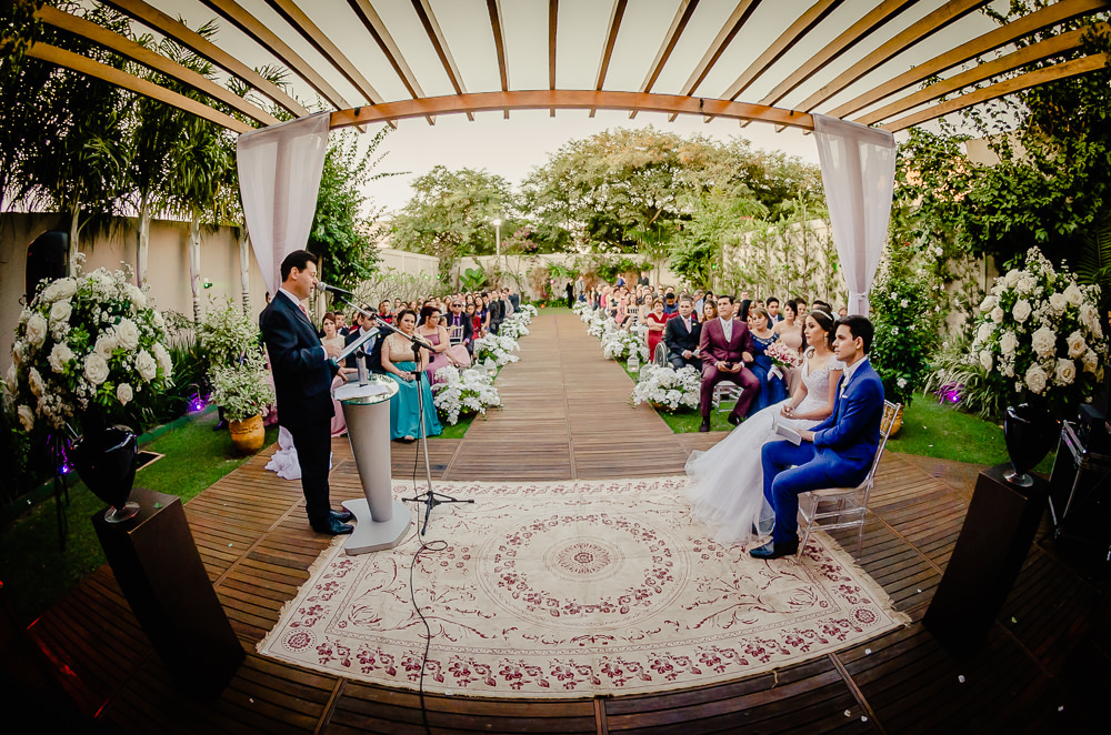 Fotografia da cerimônia de casamento com o orador do discurso de casamento e os noivos sentados prestando atenção e os convidados ao fundo. A decoração é com flores brancas e coloridas no espaço de eventos ao ar livre durante o dia e com sol em Maringá PR