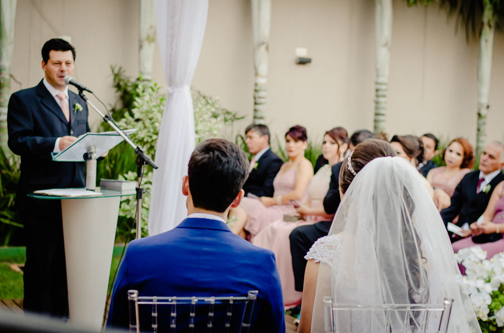 Fotografia da cerimônia de casamento com os noivos prestando atenção ao discurso de casamento e os convidados ao fundo, ela de véu branco e ele de terno azul em um espaço de eventos ao ar livre durante o dia e com sol em Maringá PR