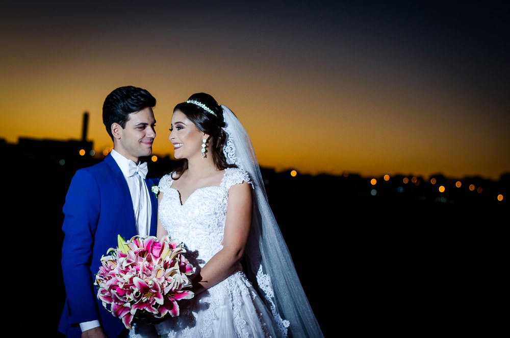 Fotografia do ensaio de casamento após a cerimonia do casal de noivos no pôr-do-sol em campo aberto, ela segura o buquê de vestido de noiva longo e branco com o véu de noiva branco e longo. O noivo de terno azul em Maringá PR