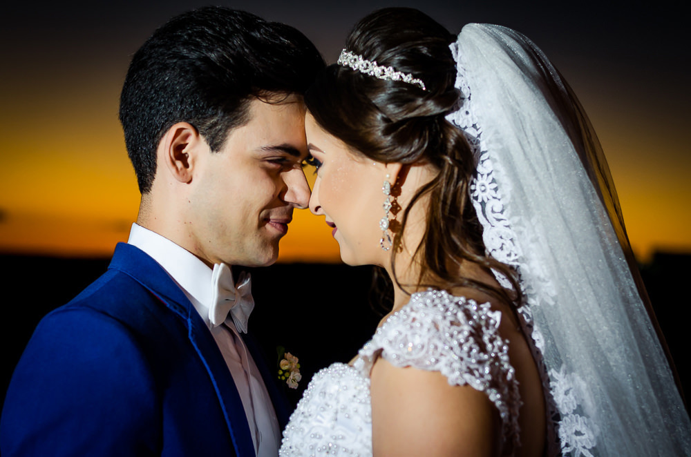 Fotografia do ensaio de casamento após a cerimonia do casal de noivos no pôr-do-sol em campo aberto, ela segura o buquê de vestido de noiva longo e branco com o véu de noiva branco e longo. O noivo de terno azul em Maringá PR