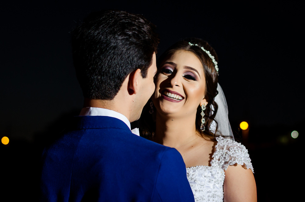 Fotografia do ensaio de casamento após a cerimonia do casal de noivos a noite em campo aberto, ela segura o buquê de vestido de noiva longo e branco com o véu de noiva branco e longo. O noivo de terno azul em Maringá PR