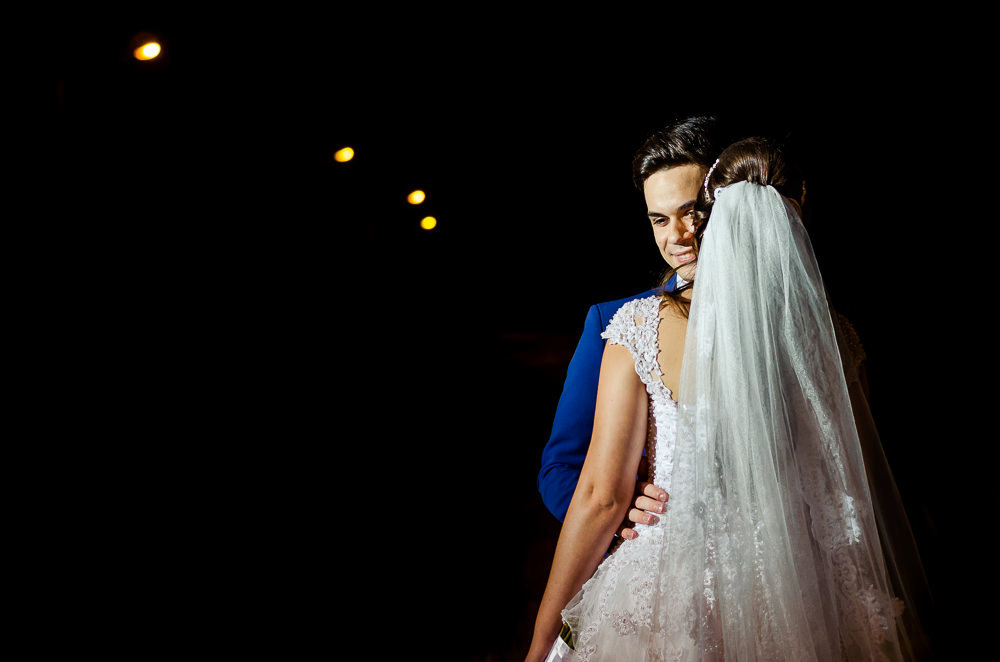 Fotografia do ensaio de casamento após a cerimonia do casal de noivos a noite em campo aberto, ela segura o buquê de vestido de noiva longo e branco com o véu de noiva branco e longo. O noivo de terno azul em Maringá PR
