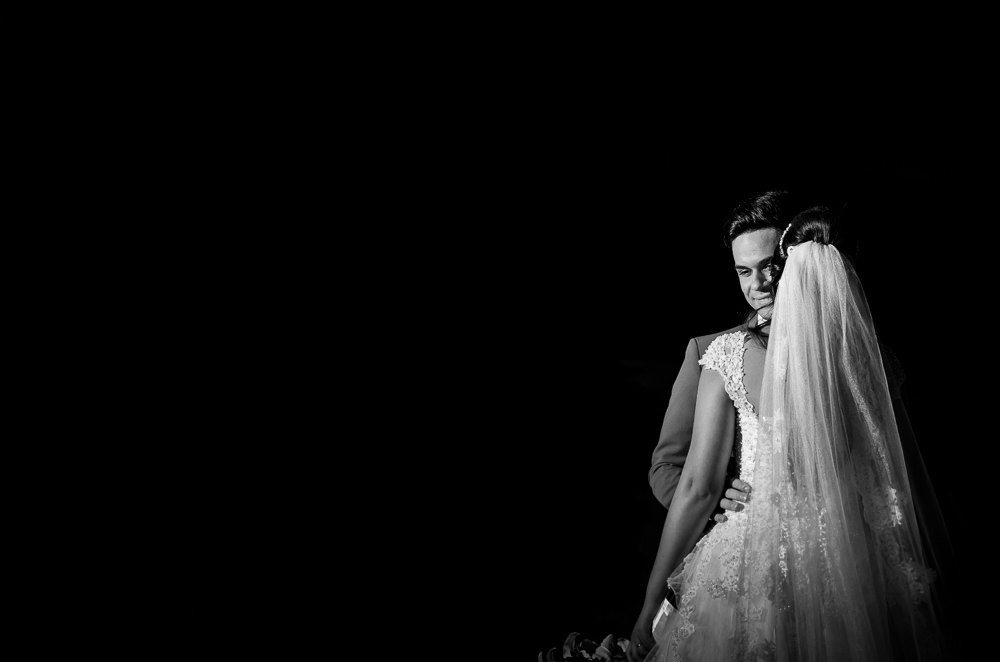 Fotografia em preto e branco, pb, do ensaio de casamento após a cerimonia do casal de noivos a noite em campo aberto, ela segura o buquê de vestido de noiva longo e branco com o véu de noiva branco e longo. O noivo de terno azul em Maringá PR