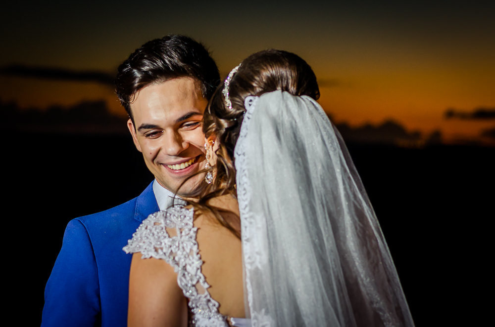 Fotografia do ensaio de casamento após a cerimonia do casal de noivos no pôr-do-sol em campo aberto, ela segura o buquê de vestido de noiva longo e branco com o véu de noiva branco e longo. O noivo de terno azul em Maringá PR
