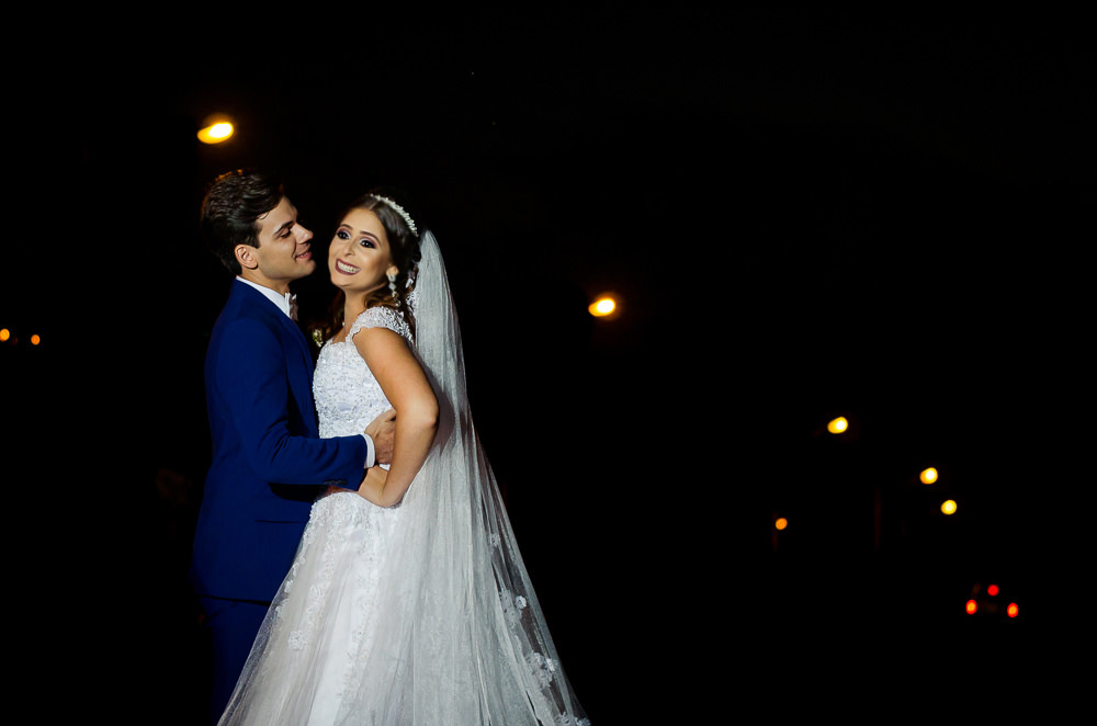 Fotografia do ensaio de casamento após a cerimonia do casal de noivos a noite em campo aberto, ela segura o buquê de vestido de noiva longo e branco com o véu de noiva branco e longo. O noivo de terno azul em Maringá PR