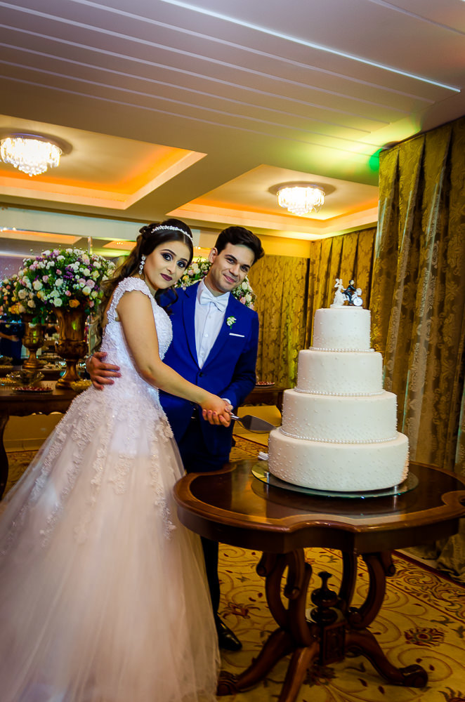 Fotografia da recepção de casamento dos noivos cortando o bolo de casamento. A noiva de vestido de noiva branco e longo com tiara no cabelo, o noivo de terno azul e gravata borboleta branca no espaço ou salão de eventos para festas em Maringá PR