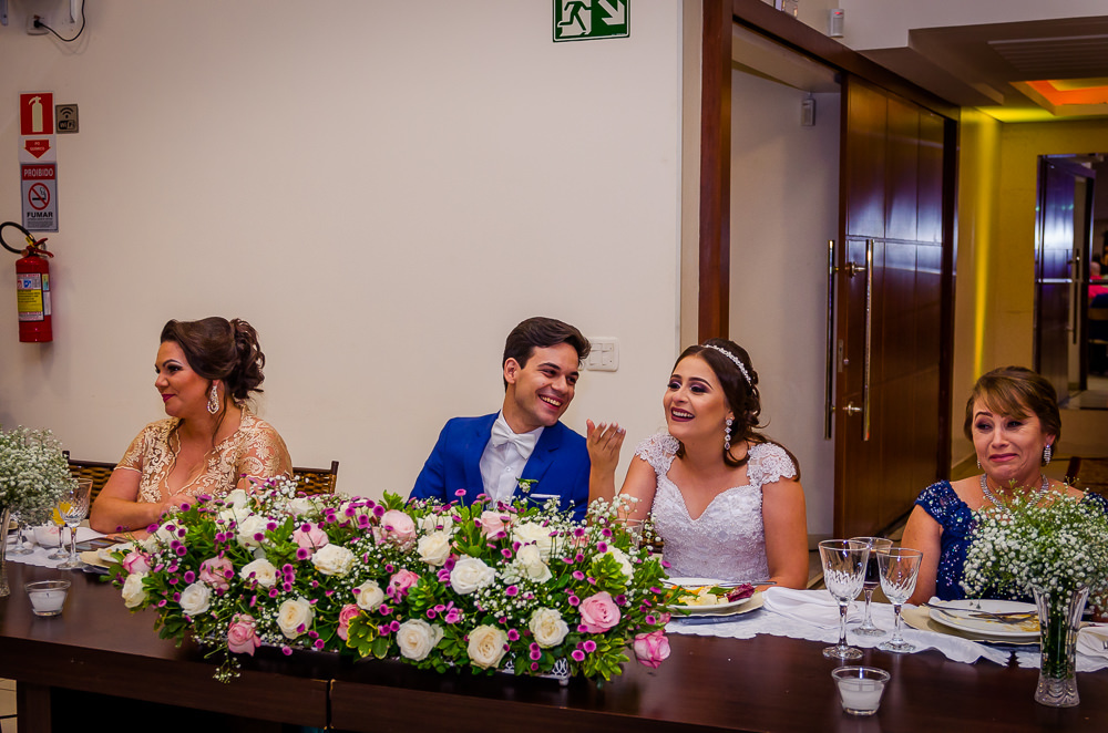 Fotografia da recepção ou festa de casamento dos noivos sentados e sorrindo e flores brandas e coloridas como decoração da mesa no espaço de eventos em Maringá PR