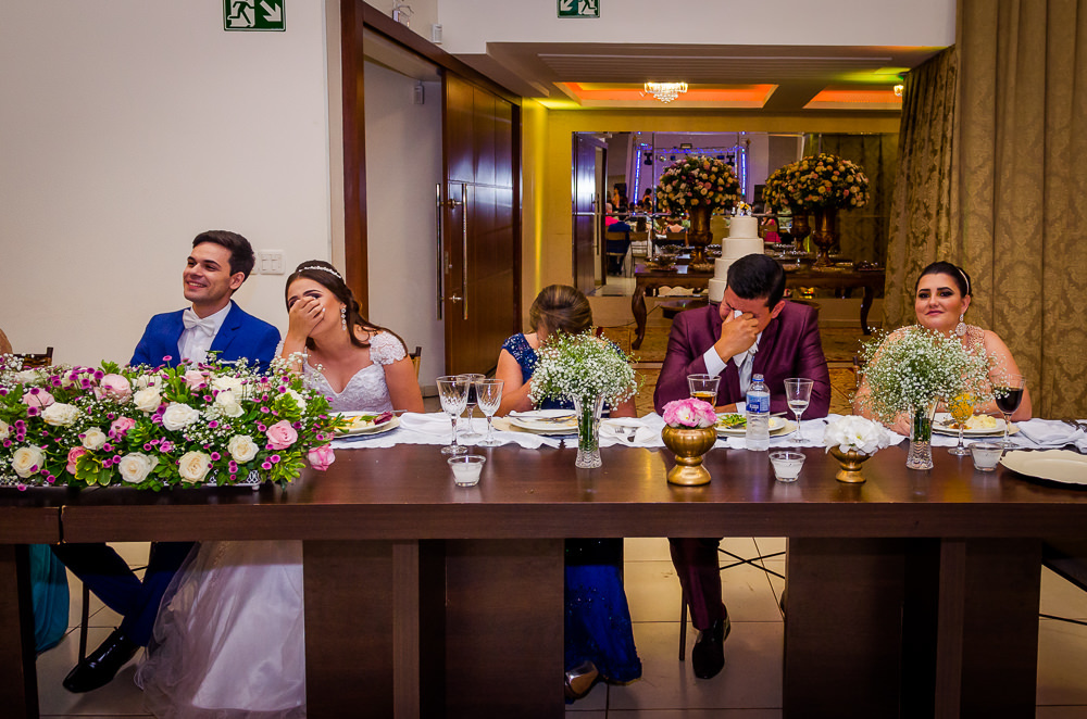Fotografia da recepção ou festa de casamento dos noivos sentados e flores brancas e coloridas como decoração da mesa. A noiva, sua mãe e seu irmão estão chorando emocionados no espaço de eventos em Maringá PR