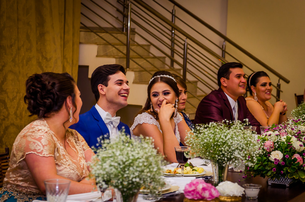 Fotografia da recepção ou festa de casamento dos noivos sentados e sorrindo e flores brancas e coloridas como decoração da mesa no espaço de eventos em Maringá PR