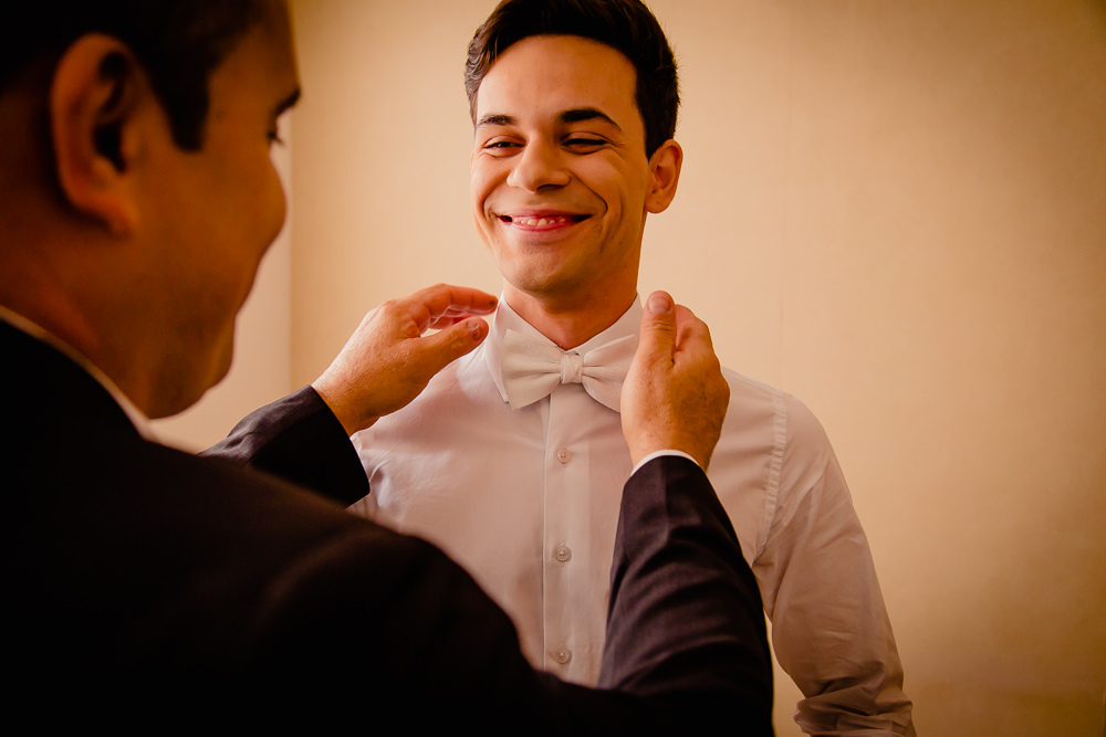 Fotografia de making of do noivo no dia do casamento com seu pai arrumando a gravata borboleta branca e usando camisa branca, o pai e o filho sorrindo em Maringá PR