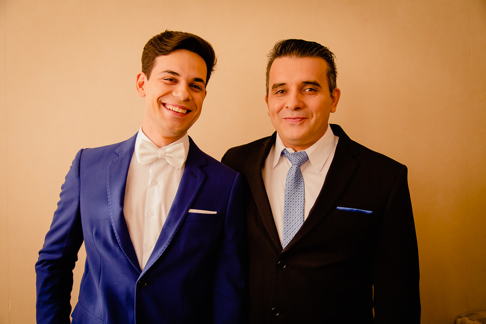 Fotografia de making of do noivo sorrindo no dia do casamento de terno azul, usando gravata borboleta branca e camisa branca junto com seu pai de terno preto e gravata azul sorrindo em Maringá PR