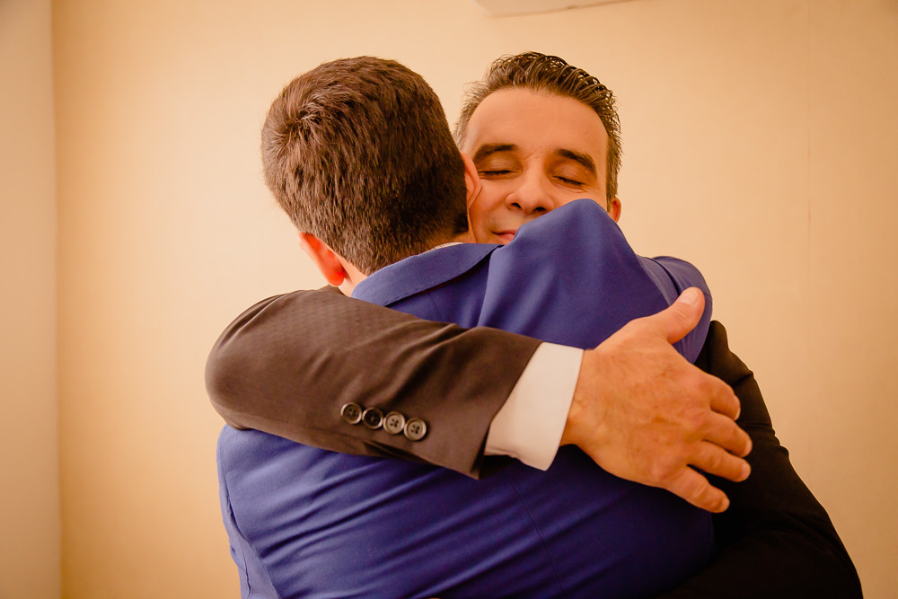 Fotografia de making of do noivo no dia do casamento de terno azul, usando gravata borboleta branca e camisa branca junto abraçando seu pai de terno preto e gravata azul sorrindo em Maringá PR