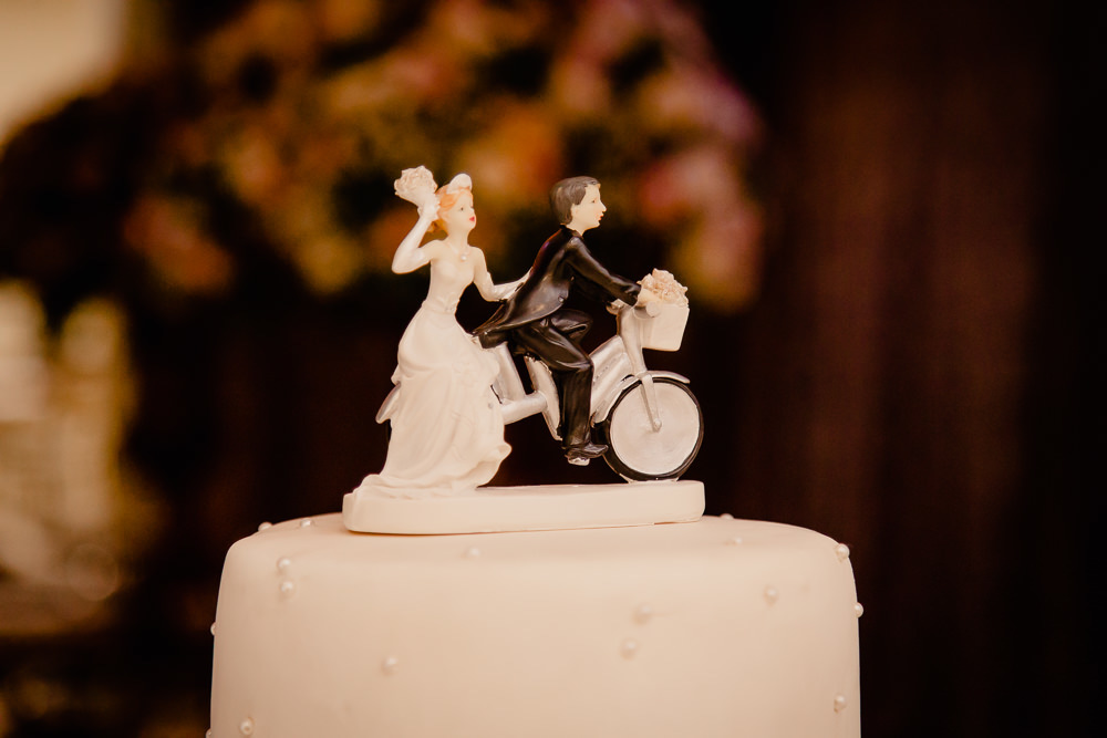 Fotografia do bolo de casamento com o noivinhos no topo do bolo, ele andando de bicicleta e ela na garupa com vestido de noiva branco em Maringá PR