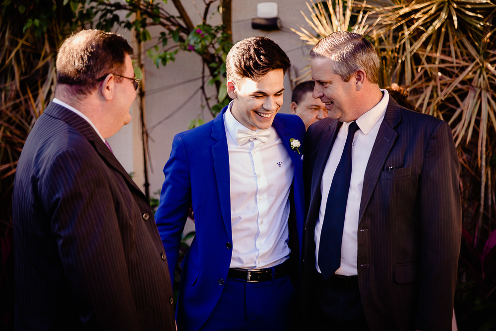 Fotografia do dia da cerimônia de casamento do noivo usanto camisa branca e terno azul com gravata borboleta branda conversando e sorrindo com amigos convidados antes da cerimônia no salão de eventos durante o dia ensolarado em Maringá PR