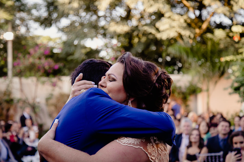 Fotografia do noivo no dia da cerimônia de casamento vestindo terno azul abraçando sua mãe emocionados. Cerimônia durante o dia a céu aberto e ensolarado no espaço de eventos em Maringá PR