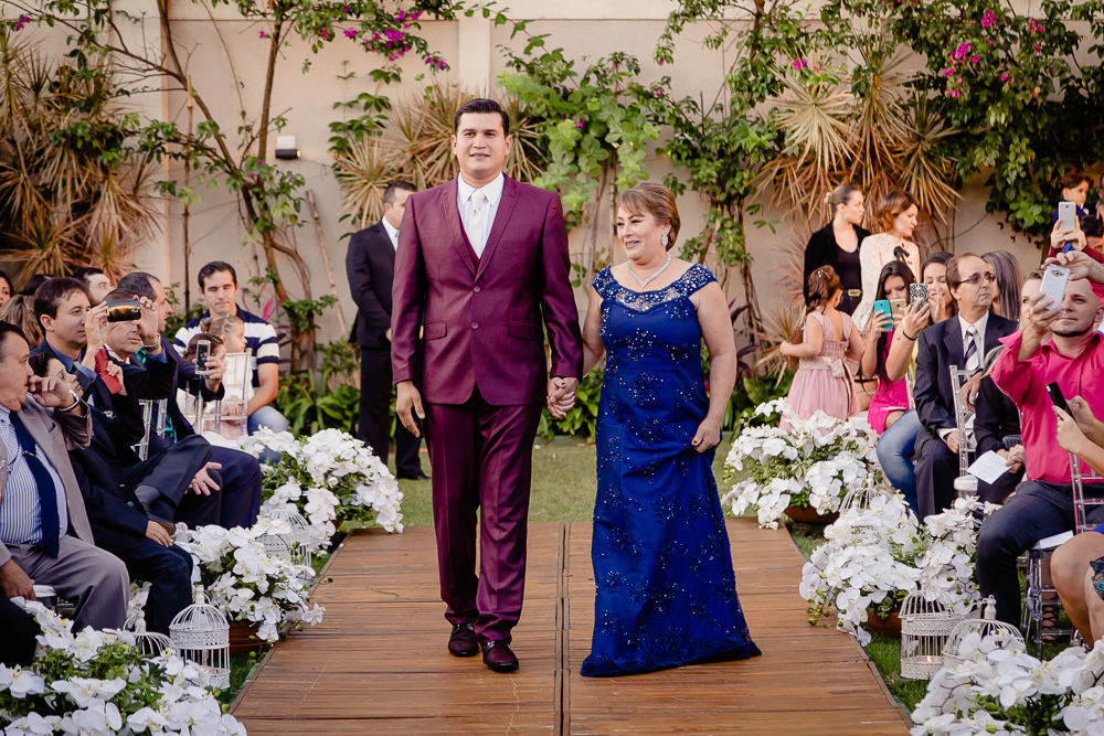 Fotografia da cerimônia de casamento durante o dia, a céu aberto e com sol no espaço de eventos. O irmão da noiva de terno roxo e gravata tradicional branca caminhando com a mãe da noiva usando vestido longo azul na cidade de Maringá PR