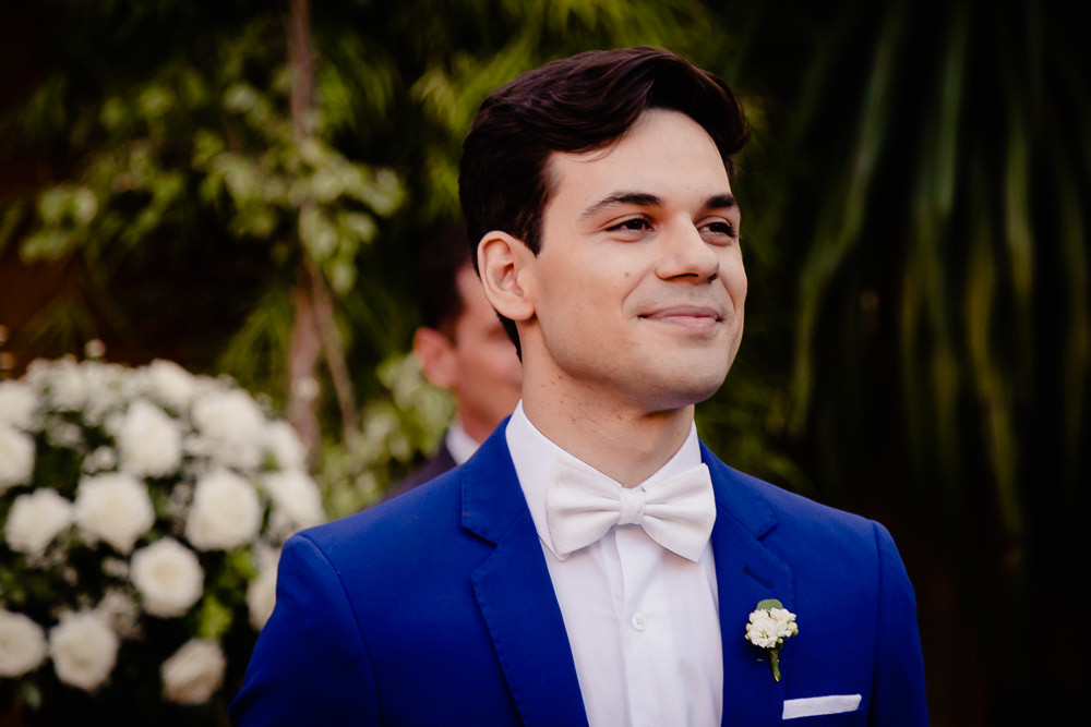 Fotografia do noivo no dia do casamento na cerimônia olhando com olhar feliz para a noiva entrando. Ele usa terno azul e gravata borboleta branca, no espaço de eventos durante o dia a céu aberto com sol em Maringá PR