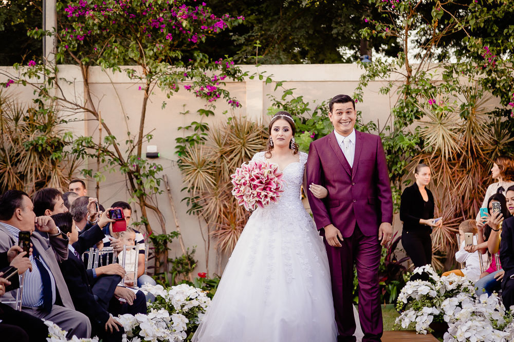 Fotografia da cerimônia de casamento com os convidados. A noiva entra com vestido longo branco e buquê de flores junto com seu irmão que sorri ao caminhar na nave e usa terno roxo no espaço de eventos durante o dia, a céu aberto com sol em Maringá PR 
