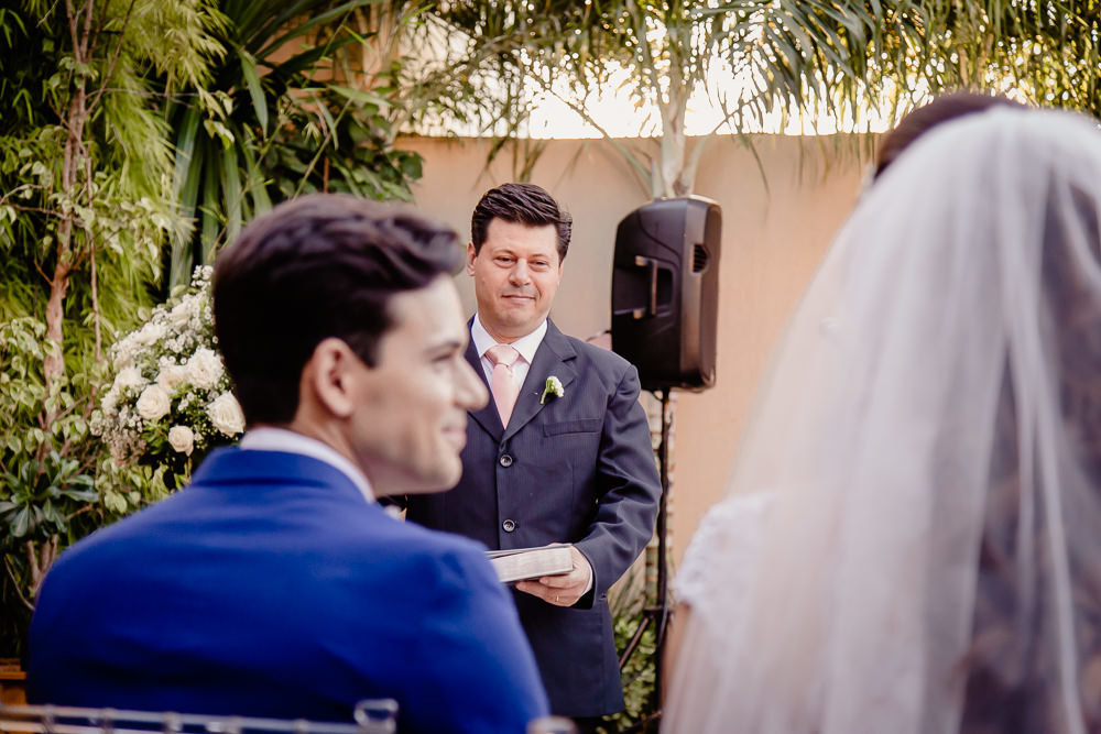 Fotografia da cerimônia de casamento do noivo usando terno azul olhando para a noiva de véu branco no espaço de eventos a céu aberto, durante o dia e com sol em Maringá PR