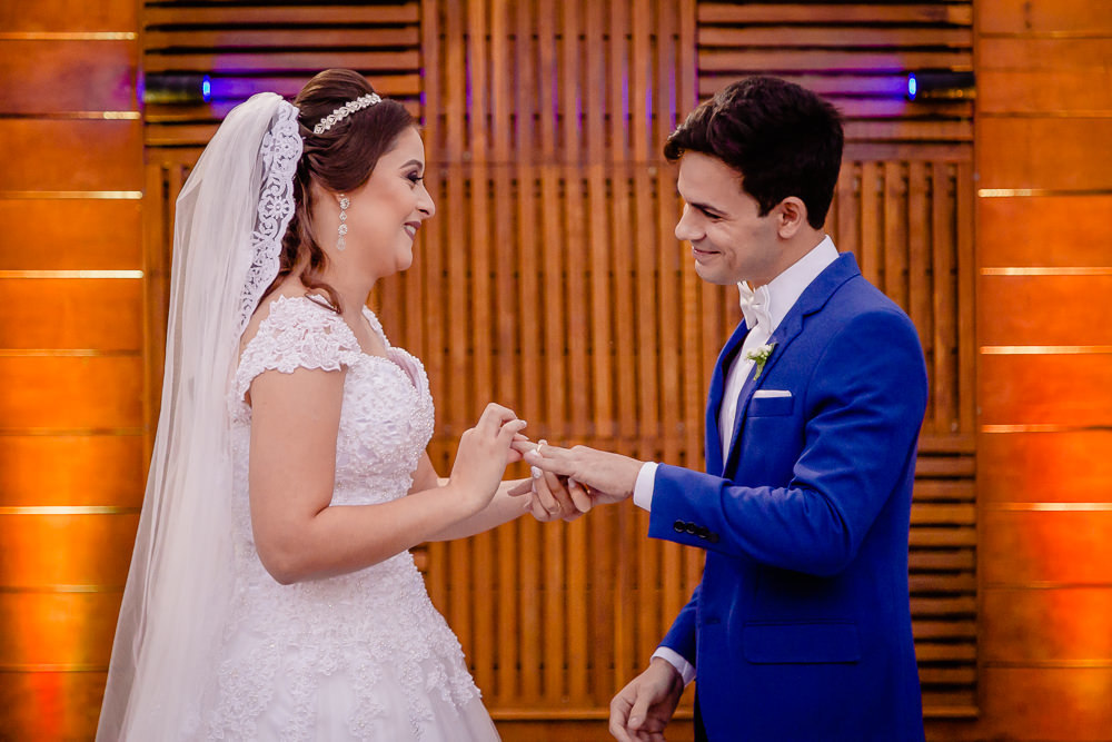Fotografia da cerimônia de casamento dos noivos trocando as alianças. A noiva de vestido de noiva branco longo e véu branco coloca a aliança na mão noivo de terno azul no espaço de eventos a céu aberto, durante o dia e ensolarado em Maringa PR