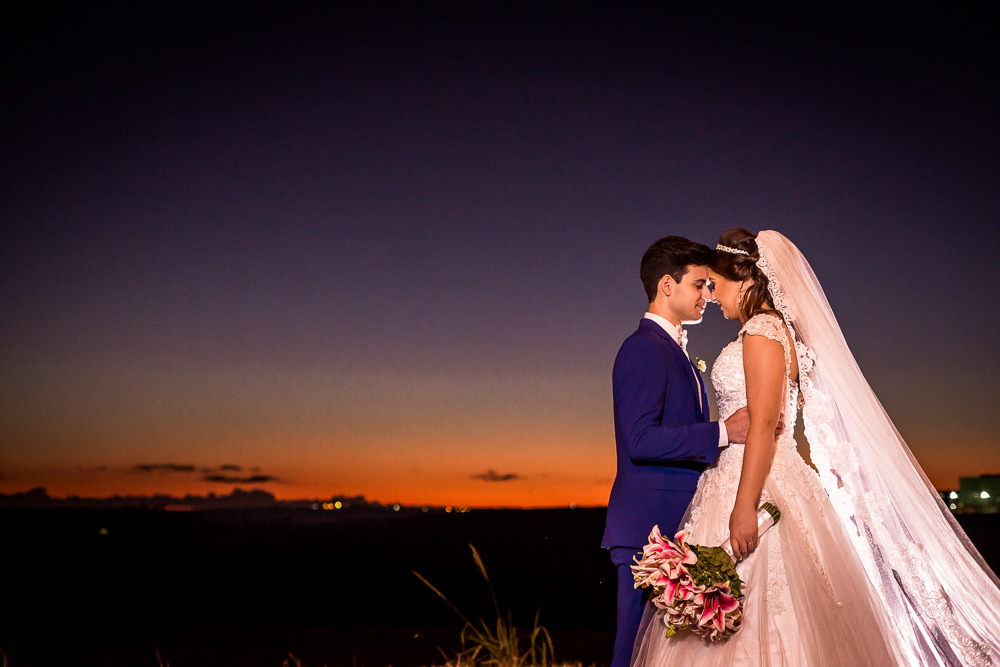 Fotografia do ensaio de casamento após a cerimonia do casal de noivos no pôr-do-sol em campo aberto, ela segura o buquê de vestido de noiva longo e branco com o véu de noiva branco e longo. O noivo de terno azul em Maringá PR