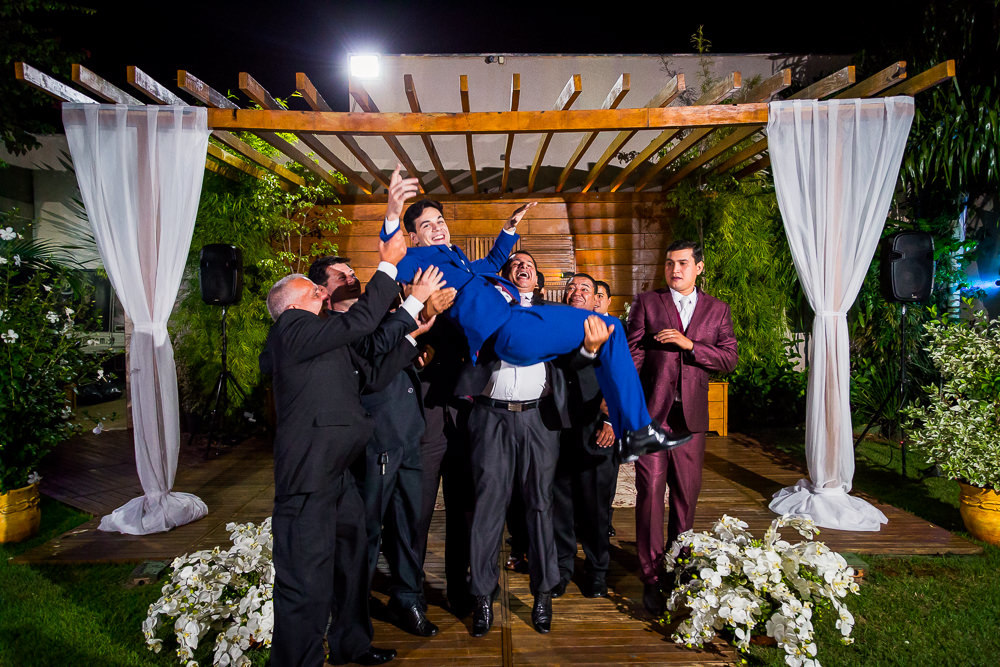 Fotografia da recepção ou festa de casamento do noivo de terno azul e gravata borboleta branca com os padrinhos lançando ou jogando o noivo para cima no espaço a céu aberto do evento em Maringá PR
