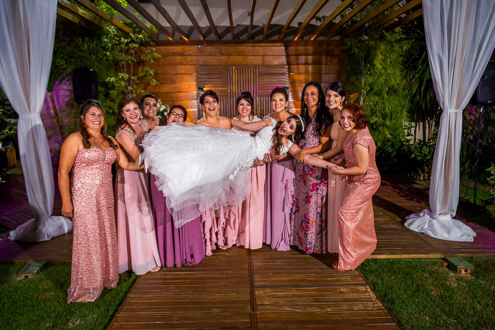 Fotografia da recepção ou festa de casamento no espaço a céu aberto da noiva usando vestido de noiva branco e longo sorrindo e as madrinhas de casamento de vestidos de tons de rosa segurando a noiva nos braços no espaço de eventos em Maringá, PR