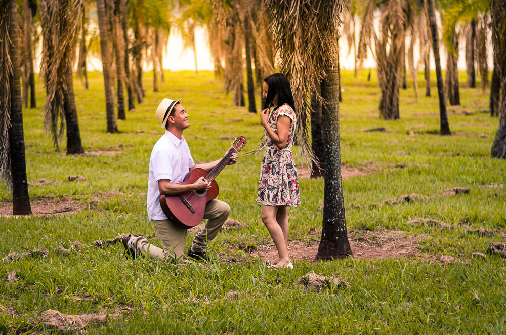 Fotografia do ensaio de pre casamento ou wedding dos noivos em um campo aberto com muitas plameiras e sol. Estão próximos, ele toca uma canção no violão para ela de joelhos. Em Araçatuba, SP