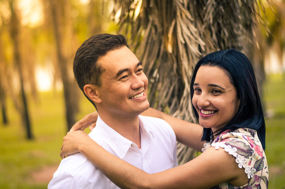 Fotografia no campo ensolarado na natureza com palmeiras ao fundo do casal de noivos no ensaio pre casamento ou wedding. Ela abraça o noivo e ambos sorriem com satisfação.