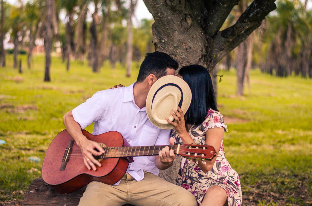 Ensaio pre casamento ou wedding do casal de noivos em Araçatuba, SP. Estão sentados, ele toca uma música no violão e ela tampa o rosto dels com um chapéu.