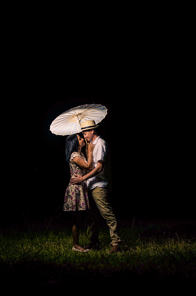 Fotografia do ensaio pre casamento ou wedding do casal de noivos em Araçatuba, SP. Está a noite estão se abraçando e se olhando com carinho, ele usa chapéu e com uma sombrinha japonesa, Wagasa