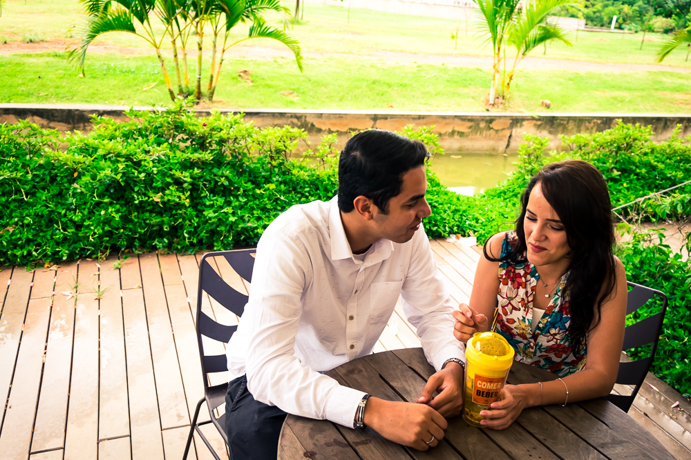 Foto do casal de noivos no ensaio pre casamento sentados a mesa conversando segurando um copo grande amarelo que tem significados especial para o casal. Ensaio no Parque Vila Lobos, Alto de Pinheiros, São Paulo, SP no dia de chuva.