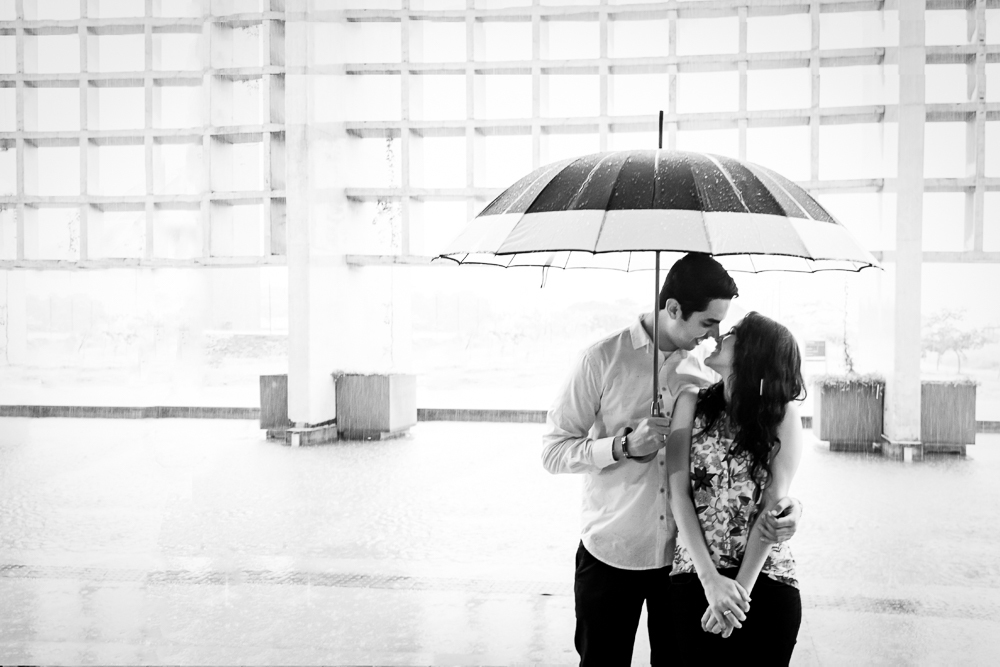 Foto  preto e branco, pb, do casal de noivo no ensaio pre casamento. Ele segura um guarda-chuva e a abraça. Ensaio no Parque Vila Lobos, Alto de Pinheiros, São Paulo, SP em uma dia de muita chuva.