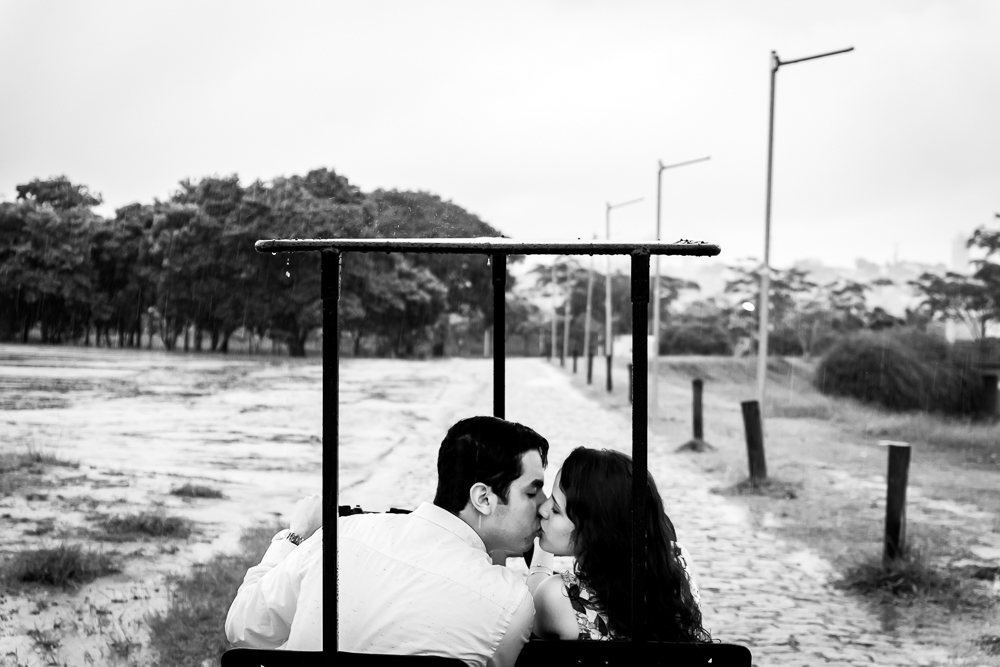 Fotografia do casal de noivos. A noiva e o noivo estão juntos pedalando a bicicleta e se beijando. Ensaio no Parque Vila Lobos, Alto de Pinheiros, São Paulo, SP em uma dia de muita chuva.