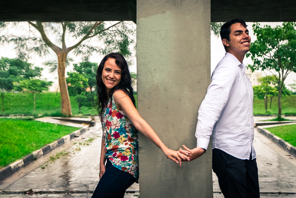 Foto do casal de noivos no ensaio fotográfico pre wedding, casamento. Eles estão em baixo de uma ponte pequena separados por uma parede grossa de concreto e apenas de mãos dadas e sorrindo felizes. Ensaio no Parque Vila Lobos, Alto de Pinheiros, São Paulo