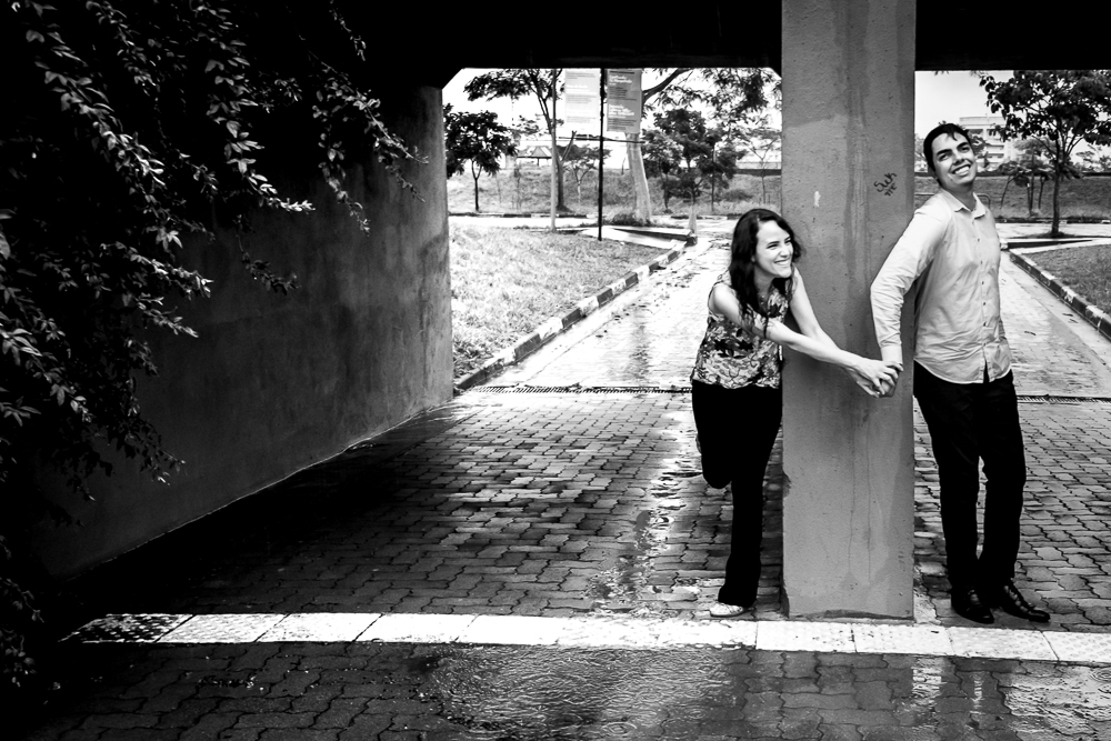 Foto do casal de noivos no ensaio fotográfico pre wedding, casamento. Eles estão em baixo de uma ponto pequena separados por uma parede grossa de concreto e apenas de mãos dadas e sorrindo felizes. Ensaio no Parque Vila Lobos, Alto de Pinheiros, São Paulo