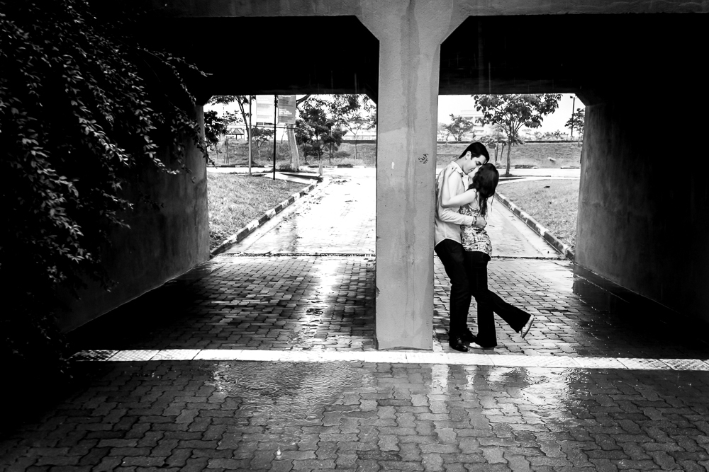 Foto do casal de noivos no ensaio fotográfico pre wedding, casamento. Eles estão em baixo de uma ponte se abraçando e beijando do lado direito. Ensaio no Parque Vila Lobos, Alto de Pinheiros, São Paulo