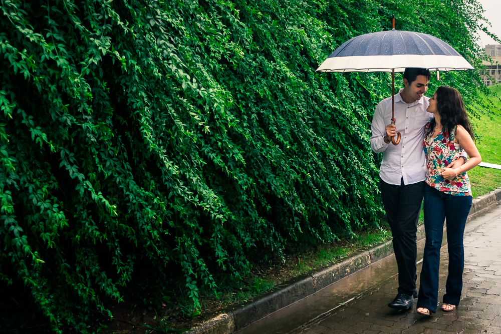 Foto do casal de noivos no ensaio pre casamento caminhando de guarda-chuva na chuva se abraçando e sorrindo. Ensaio fotográfico no Parque Vila Lobos, Alto de Pinheiros, São Paulo, SP em uma dia de muita chuva.