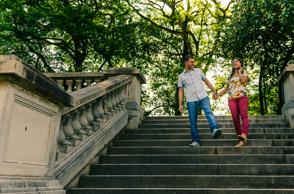 Foto do ensaio pre wedding, casamento, do casal de noivos descendo de mãos dadas uma escadaria com árvores ao fundo num dia de sol. O noivo e a noiva estão no Parque do Ipiranga, Bairro Ipiranga, São Paulo, Brasil.