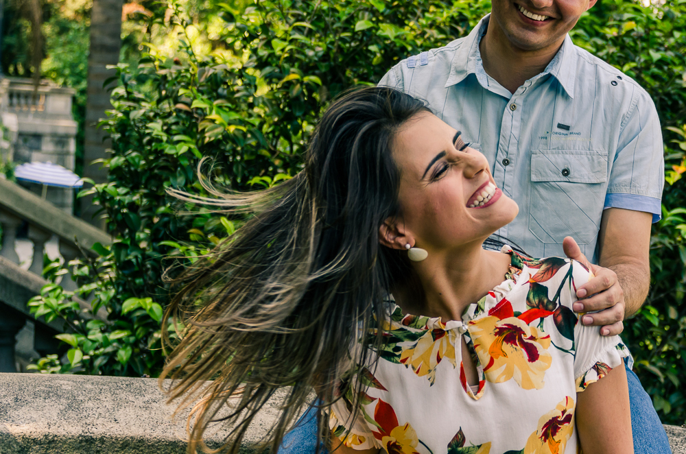 Foto do Ensaio fotográfico pré casamento, wedding do casal de noivos, ela dá risada enquanto mexe seus cabelos soltos. O noivo e a noiva estão no Parque do Ipiranga, Bairro Ipiranga, São Paulo, Brasil.