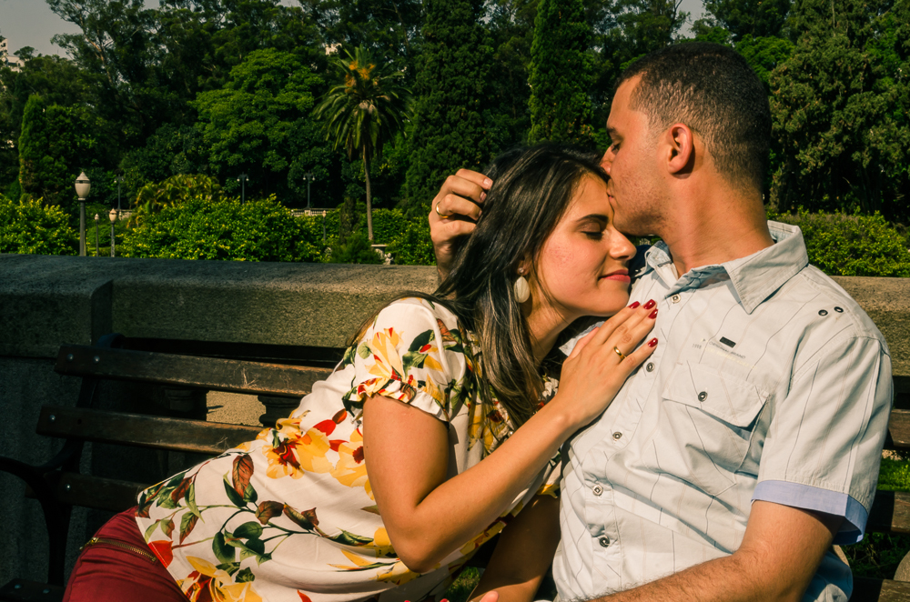 Foto do ensaio fotográfico pre casamento, wedding do casal de noivos sentados num banco, ele baija com carinho a teta da noiva. O noivo e a noiva estão no Parque do Ipiranga, Bairro Ipiranga, São Paulo, Brasil.