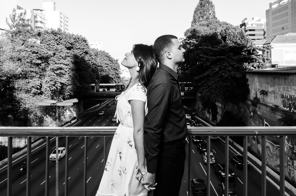 Foto do Ensaio pre Casamento, Wedding do casal de noivos de mãos dadas em cima da ponte com avenida movimentada ao fundo num dia de sol. O noivo e a noiva estão no Bairro Liberdade, São Paulo, Brasil.