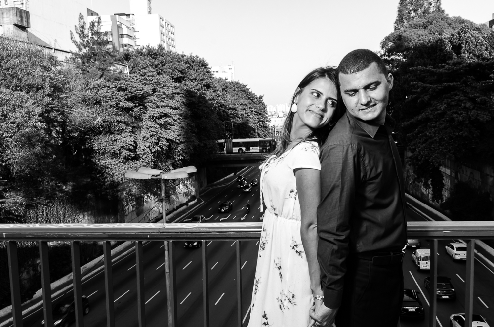 Foto do Ensaio pre Casamento, Wedding do casal de noivos de mãos dadas em cima da ponte com avenida movimentada ao fundo num dia de sol. O noivo e a noiva estão no Bairro Liberdade, São Paulo, Brasil.