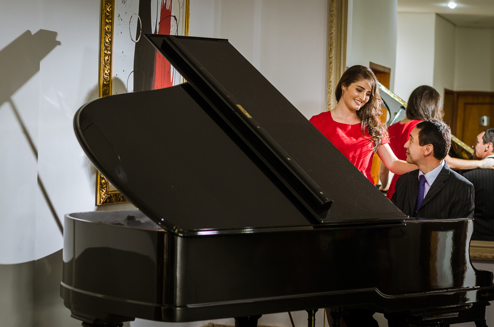 Foto do ensaio pre wedding, casamento, do casal de noivos. Estão ao piano de cauda, o noivo de traje completo, terno e gravata e a noiva de vestido vermelho no Hotel Capo Zorial no Recanto do Maestro em São João do Polêsine, RS, próximo de Santa Maria, RS