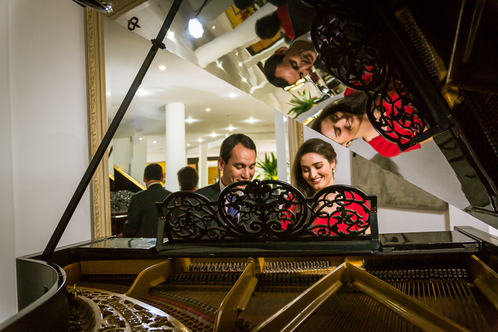 Foto do ensaio pre wedding, casamento, do casal de noivos. Estão ao piano de cauda, o noivo de traje completo, terno e gravata e a noiva de vestido vermelho no Hotel Capo Zorial no Recanto do Maestro em São João do Polêsine, RS, próximo de Santa Maria, RS