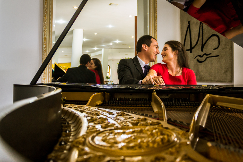 Foto do ensaio pre wedding, casamento, do casal de noivos. Estão ao piano de cauda, o noivo de traje completo, terno e gravata e a noiva de vestido vermelho no Hotel Capo Zorial no Recanto do Maestro em São João do Polêsine, RS, próximo de Santa Maria, RS