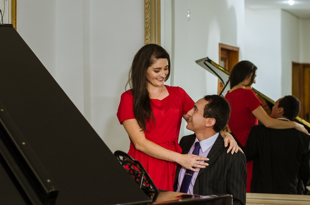 Foto do ensaio pre wedding, casamento, do casal de noivos. Estão ao piano de cauda, o noivo de traje completo, terno e gravata e a noiva de vestido vermelho no Hotel Capo Zorial no Recanto do Maestro em São João do Polêsine, RS, próximo de Santa Maria, RS