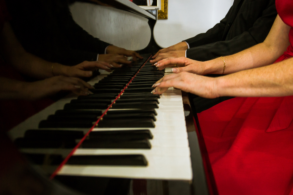 Foto do ensaio pre wedding, casamento, do casal de noivos. Estão ao piano de cauda, o noivo de traje completo, terno e gravata e a noiva de vestido vermelho no Hotel Capo Zorial no Recanto do Maestro em São João do Polêsine, RS, próximo de Santa Maria, RS