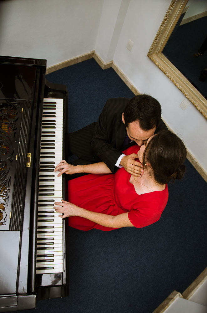 Foto do ensaio pre wedding, casamento, do casal de noivos. Estão ao piano de cauda, o noivo de traje completo, terno e gravata e a noiva de vestido vermelho no Hotel Capo Zorial no Recanto do Maestro em São João do Polêsine, RS, próximo de Santa Maria, RS
