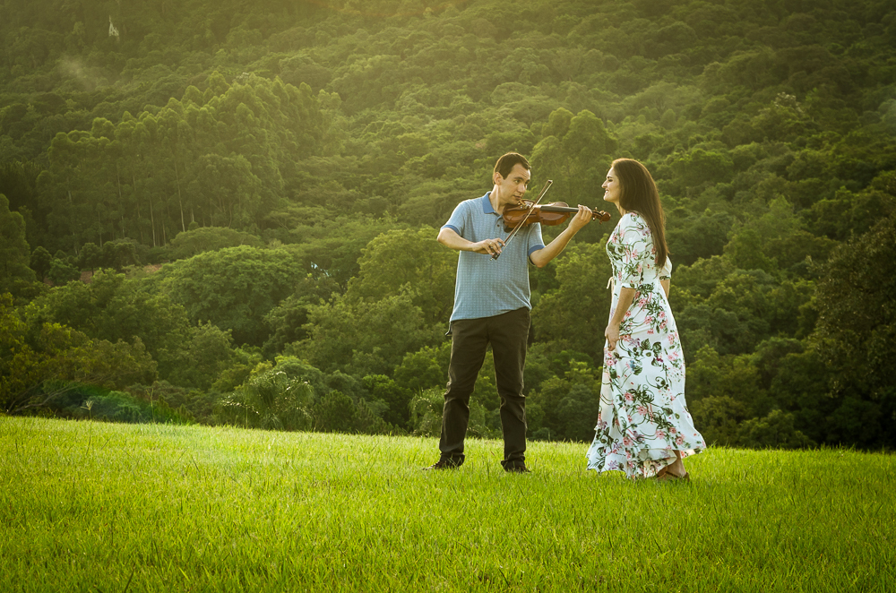 Foto do ensaio pre wedding, casamento, do casal ao ar livre com sol e céu azul tocando violino no Hotel Capo Zorial com piscina e monte ao fundo no Recanto do Maestro em São João do Polêsine, RS, próximo de Santa Maria, RS